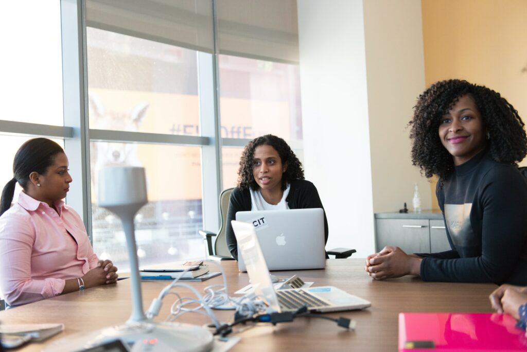 Three people sat around a table in a hybrid meeting space. One with a laptop and the others discussing.