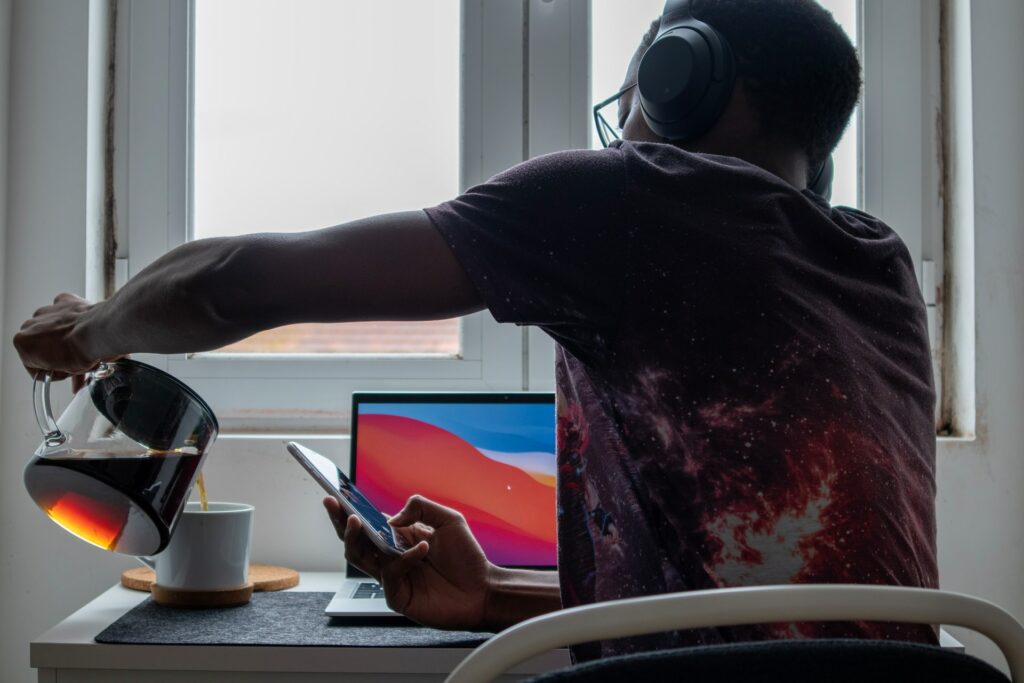 Someone sat at a desk in front of a laptop pouring coffee in to a mug while they use their phone and employing time management to create a work schedule.